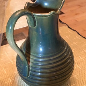 May include: A ceramic pitcher with a dark teal and green glaze. The pitcher has a curved handle and a ruffled rim. Horizontal ridges encircle the body of the pitcher. The pitcher is sitting on a woven mat.