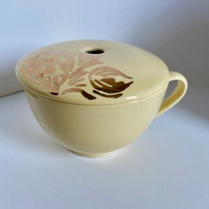 May include: A cream-colored ceramic bowl with a lid. The lid has a gold and pink floral design. The bowl has a handle on the side.