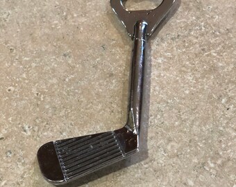 May include: A chrome-finished bottle opener shaped like a golf club. The opener has a key-shaped top with a bottle cap remover and a golf club head at the bottom. The club head has horizontal grooves.