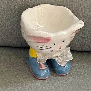 Könnte beinhalten: Ein weißer Keramik-Eierbecher in Hasenform mit rosa Innenohren, schwarzen Schnurrhaaren und einer weißen Schleife. Der Hase trägt gelbe Kleidung und blaue Schuhe mit rosa Sohlen.