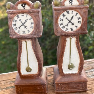 May include: Two brown ceramic grandfather clock figurines. Each clock has a white clock face with black Roman numerals and black hands. The clock bodies are brown with white and green accents. The figurines are approximately 6 inches tall.