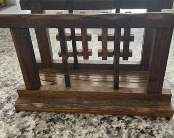 May include: A dark brown wooden magazine rack with a rectangular base and a slatted design. The rack features vertical supports and a decorative grid pattern, showcasing a rustic aesthetic. The wood grain is visible, adding to its natural appeal.