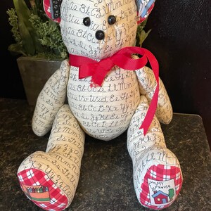 Puede incluir: Un oso de peluche con una cinta roja alrededor del cuello. El oso está cubierto de tela con letras y números escritos a mano. Las orejas del oso están decoradas con cuadrados de tela que dicen "MATH" y "ABCs". Los pies del oso están decorados con cuadrados de tela que dicen "Mi Casa" y "Crayones".