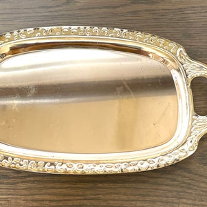 Puede incluir: Una bandeja plateada rectangular con asas ornamentadas y un borde decorativo. La bandeja tiene una superficie pulida y reflectante, ideal para servir alimentos o exhibir objetos. Las asas son curvas y añaden elegancia al diseño.