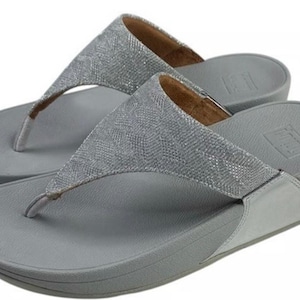 以下が含まれることがあります： グレーのFitFlopビーチサンダルのペア。フットベッドとソールは無地のグレーで、ストラップは質感のあるキラキラ光るシルバー素材です。ビーチサンダルにはわずかなウェッジヒールが付いています。