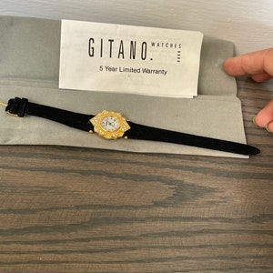 May include: A gold-tone wristwatch with a white face and diamond-like accents around the bezel. The watch is in a black velvet box with a warranty card that reads "Gitano Watches 5 Year Limited Warranty".