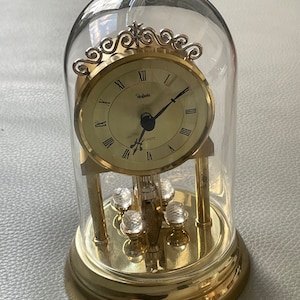 May include: A gold-toned clock with a glass dome, featuring Roman numeral markings and the word "Perfecta" on the face. The clock is on a round base with a pendulum.