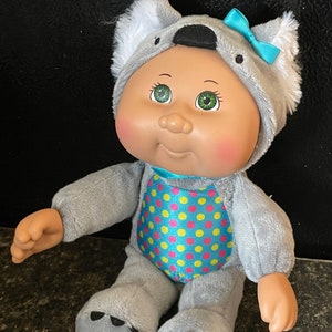 May include: A plush Cabbage Patch Kids doll wearing a gray koala costume with a blue bow. The doll has green eyes, rosy cheeks, and a polka dot pattern on the costume.