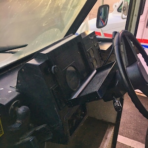 May include: Interior view of a delivery truck cab. The dashboard is black with various gauges and a fold-down tray. A black steering wheel is on the right. The windshield is visible, and a side mirror reflects another truck.