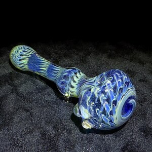 May include: A blue and white glass pipe with a swirling pattern. The pipe has a rounded bowl and a long, cylindrical stem. The colors are a mix of light blue, dark blue, and white, creating a marbled effect. The pipe is made of glass.