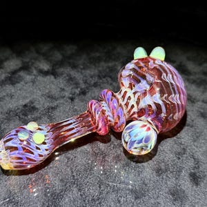 May include: A glass pipe featuring a translucent reddish-brown base with intricate white and iridescent blue swirling patterns. The piece includes a rounded bowl, a textured stem, and three small, pale yellow circular glass accents on the exterior.