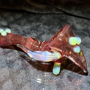 May include: A translucent pink glass pipe featuring a spiraled stem and a wide bowl. The piece is decorated with iridescent white and blue glass accents, including small spherical beads on the bowl and stem, and a curved wing-like protrusion.