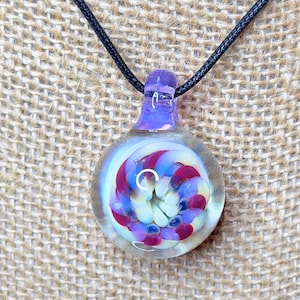 May include: A clear glass pendant with a spiral design of red, white, and blue colors. The pendant is attached to a black cord necklace. The top of the pendant is a translucent purple color.