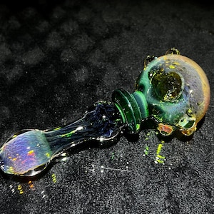 May include: A green and iridescent glass pipe with a round bowl and a long, thin stem. The bowl has a textured surface and the stem is clear with a slight iridescent sheen.
