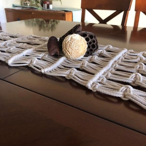 May include: A beige macrame table runner with a knotted design, placed on a dark wood table with decorative dried flower arrangements.