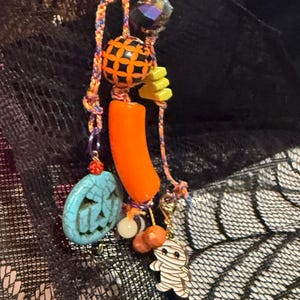 May include: A decorative charm accessory featuring a multi-colored braided cord adorned with various beads, including a turquoise jack-o'-lantern, a bright orange cylindrical bead, a yellow geometric shape, and a white mummy character charm.