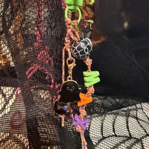 May include: A keychain featuring a bright green plastic clasp attached to a multicolored braided cord. The cord is decorated with a black ghost charm, a black bead with a white spiderweb pattern, and green, orange, and purple letter beads.