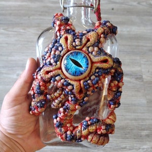 May include: A clear glass bottle with a decorative, multi-colored, tentacled design. The design features a central blue eye with a detailed iris, surrounded by red, blue, and gold textured elements. The bottle has a white stopper and a red braided cord.