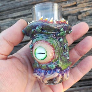 May include: A shot glass with a monster-themed design. The glass is clear, and the base is sculpted with a green, purple, and red creature design, featuring a large eye and sharp teeth. The shot glass is held in a hand.