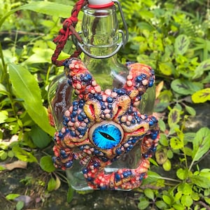 May include: A clear glass bottle with a swing-top stopper and a braided cord, featuring a sculpted, multi-tentacled creature attachment. The creature has a textured, bumpy surface in shades of red, orange, and blue, centered by a single, large, bright blue eye with a black vertical pupil.