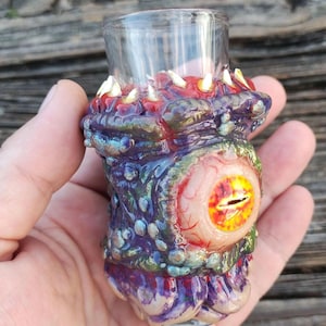 May include: A clear glass shot glass with a detailed, sculpted base. The base is painted with iridescent blues, purples, and greens, featuring a large, detailed eye and a mouth with sharp teeth. The shot glass is held in a hand.