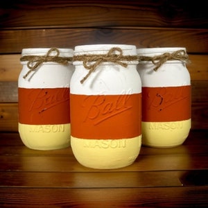 Può includere: Tre barattoli di vetro dipinti per assomigliare a caramelle di mais. I barattoli sono bianchi, arancioni e gialli. Ogni barattolo ha un fiocco di spago marrone attorno alla parte superiore e le parole "Ball Mason" stampate sul lato.