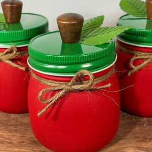 K&ouml;nnte beinhalten: Drei rote Einmachgl&auml;ser mit gr&uuml;nen Deckeln und Holzgriffen. Jedes Glas hat ein gr&uuml;nes Blatt und eine braune Kordelschleife.