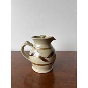 May include: A ceramic pitcher with a lid, featuring a speckled light beige glaze and a hand-painted brown leaf pattern encircling the body. The vessel includes a curved handle, a flared rim with a small pouring spout, and a circular lid.