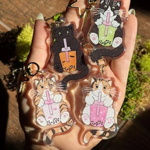 May include: Four clear acrylic cat keychains. Each keychain features a different cat design, holding a bubble tea drink. The cats are black, calico, and tabby, with gold star and round key rings.