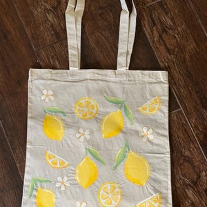 May include: A beige tote bag with a lemon design. The bag features yellow lemons, lemon slices, green leaves, and white flowers. The tote bag has two beige handles.