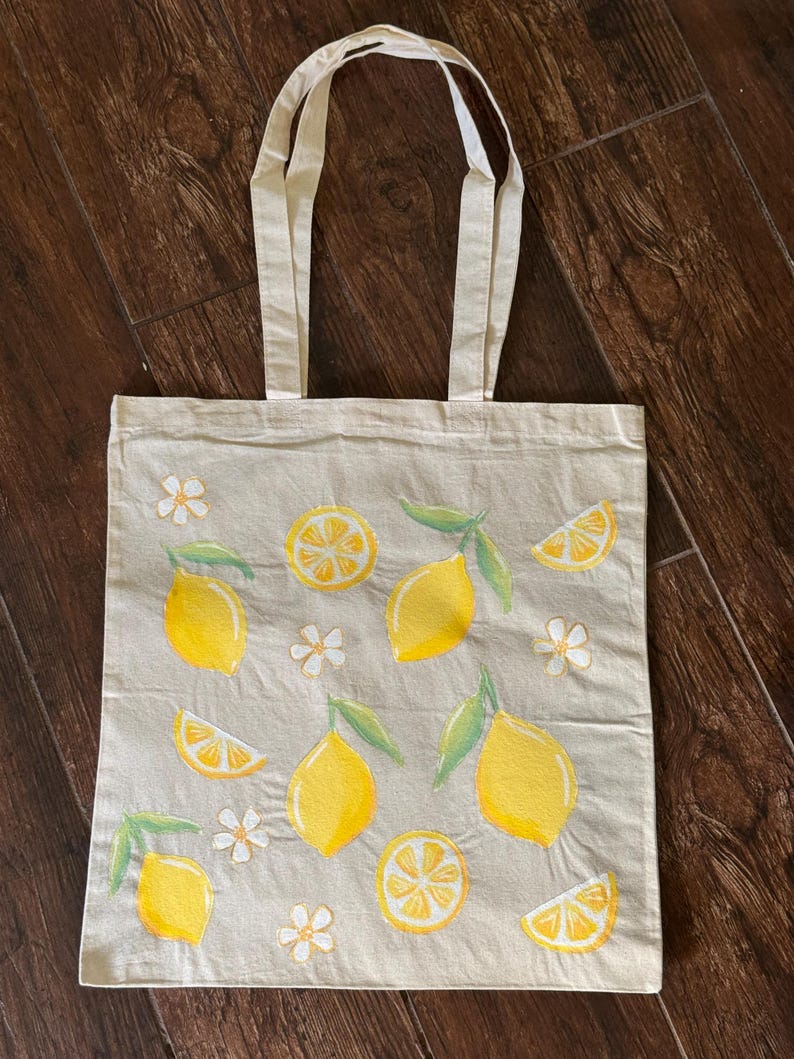 May include: A beige tote bag with a lemon design. The bag features yellow lemons, lemon slices, green leaves, and white flowers. The tote bag has two beige handles.
