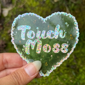 Könnte beinhalten: Ein herzförmiger Aufkleber mit grünem Hintergrund und schimmerndem Glitzer. Der Aufkleber zeigt den Text "Touch Moss" in einer verspielten Schrift.