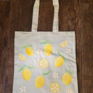 May include: A beige canvas tote bag featuring a lemon design. The bag has yellow lemons with green leaves and white flowers. The tote bag has two beige handles.
