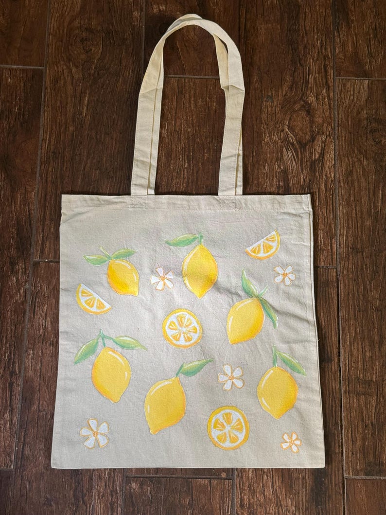 May include: A beige canvas tote bag featuring a lemon design. The bag has yellow lemons with green leaves and white flowers. The tote bag has two beige handles.