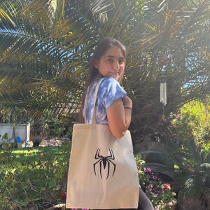 Puede incluir: Una persona lleva una bolsa de tela blanca con un logotipo de Spiderman negro. La bolsa est&aacute; hecha de lona y tiene una correa larga.