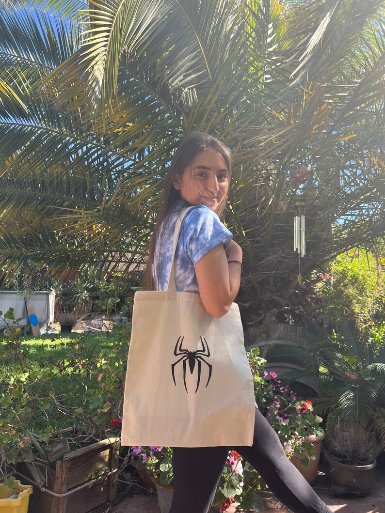 Puede incluir: Una persona lleva una bolsa de tela blanca con un logotipo de Spiderman negro. La bolsa est&aacute; hecha de lona y tiene una correa larga.