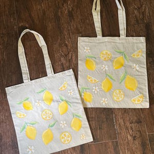 May include: Two beige tote bags with yellow lemons, lemon slices, green leaves, and white flowers. The bags have matching beige handles and are made of a light-colored fabric.