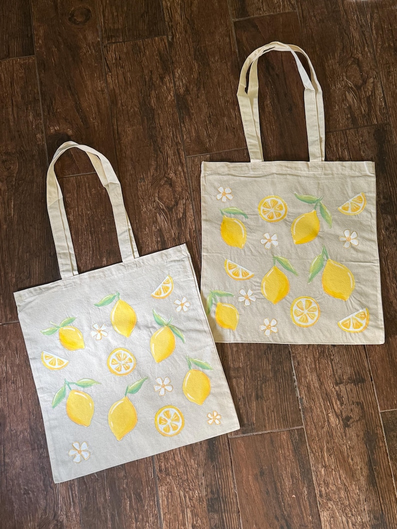 May include: Two beige tote bags with yellow lemons, lemon slices, green leaves, and white flowers. The bags have matching beige handles and are made of a light-colored fabric.