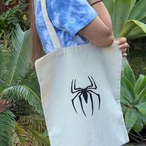 Puede incluir: Una bolsa de lona blanca con un gr&aacute;fico de silueta de Spiderman negro.