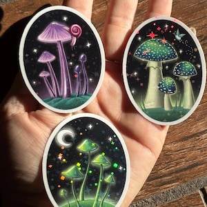 May include: Three oval stickers featuring mushroom illustrations. One sticker shows purple mushrooms with a snail, another has green and white mushrooms, and the last has green mushrooms with a crescent moon and stars. All have a black background.
