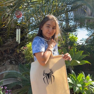 Puede incluir: Una persona sostiene una bolsa de lona beige con un gr&aacute;fico de ara&ntilde;a negra. La bolsa tiene una correa larga y est&aacute; hecha de lona.