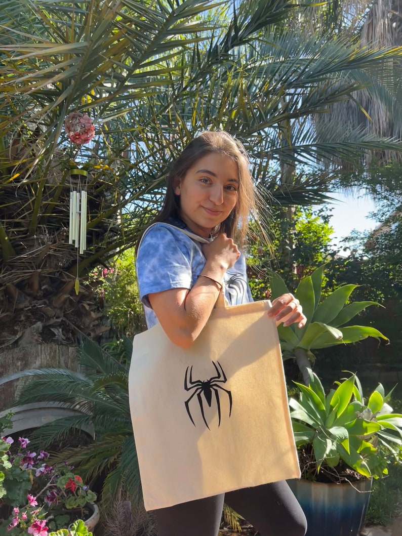 Puede incluir: Una persona sostiene una bolsa de lona beige con un gr&aacute;fico de ara&ntilde;a negra. La bolsa tiene una correa larga y est&aacute; hecha de lona.