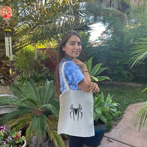 Puede incluir: Una bolsa de tela blanca con un logotipo de Spiderman negro. La bolsa tiene dos asas y la est&aacute; sujetando una persona.