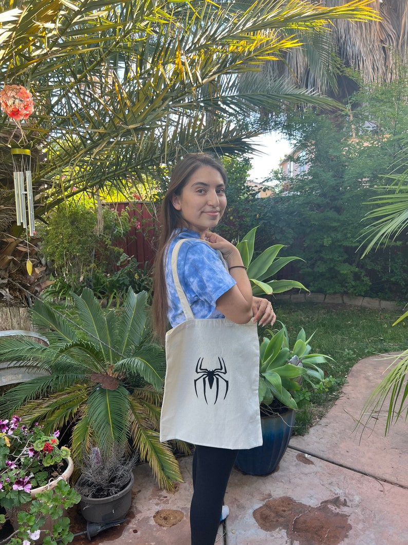 Puede incluir: Una bolsa de tela blanca con un logotipo de Spiderman negro. La bolsa tiene dos asas y la est&aacute; sujetando una persona.