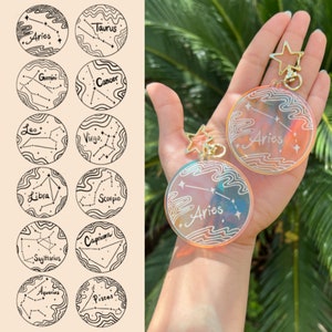 May include: Two iridescent clear keychain charms with gold star charms and rings. Each charm features a white outline of the zodiac constellation for Aries with the word "Aries" written inside.