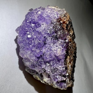 Pode incluir: Um grande aglomerado de cristais de ametista roxa com uma textura áspera e natural. O cristal é de cor roxa intensa e tem uma superfície brilhante e vítrea.