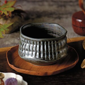 Puede incluir: Un conjunto tradicional de ceremonia del té japonesa con un cuenco de té de cerámica gris oscuro, una bandeja de madera, un batidor de bambú, una cuchara de madera y un pequeño plato blanco con un dulce.