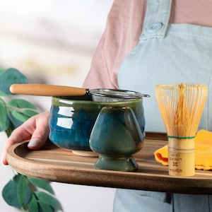 Puede incluir: Una bandeja de madera contiene un juego de té matcha. El juego incluye un cuenco de cerámica azul y verde con un colador con mango de madera, una pequeña taza de té de cerámica y un batidor de bambú. Un paño amarillo y una cuchara de bambú también están en la bandeja.