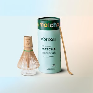 May include: A green and white cylindrical container with the text "aprika matcha master kit" and "100 prongs" on it. A bamboo whisk and a bamboo matcha scoop are next to the container.