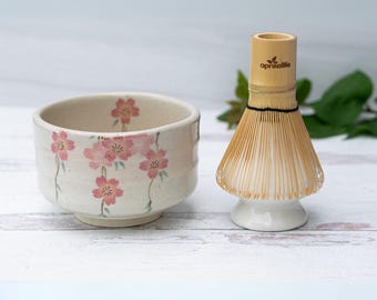Puede incluir: Un cuenco de matcha de cerámica blanca con un diseño floral rosa y un batidor de matcha de bambú con una base de cerámica blanca. El batidor tiene el texto "aprikaliife" impreso en el mango de bambú.